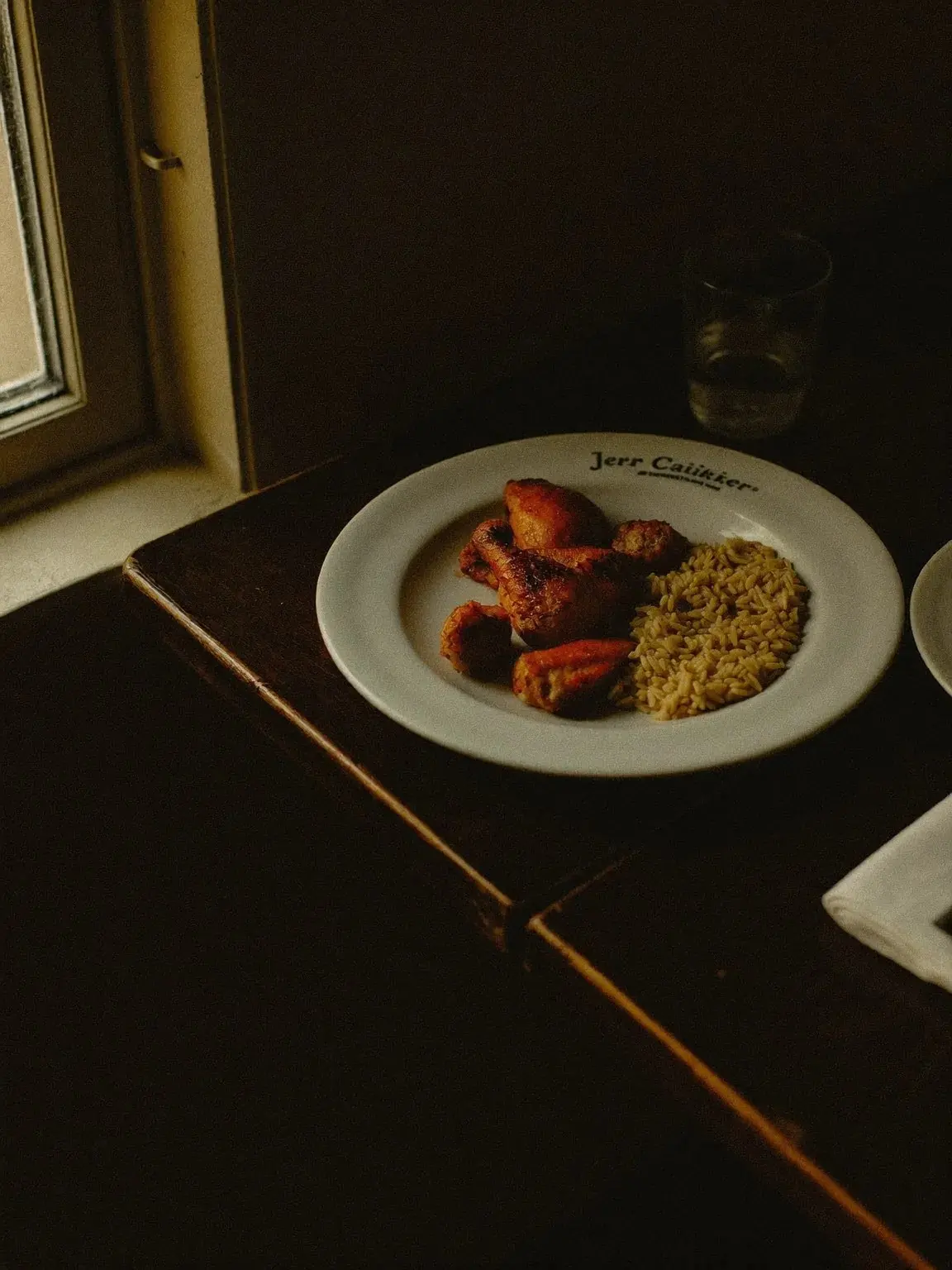 A close-up corner of a laid table with a plate of jerk chicken, rice and peas